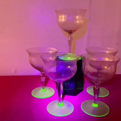 Set of five vintage Depression-era glass cocktail stems illuminated under blacklight to display green uranium fluorescence.