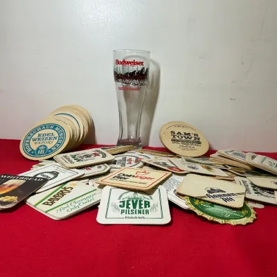 Collection of vintage assorted beer coasters featuring various brands and logos, displayed with a Budweiser Clydesdale pilsner glass on a...