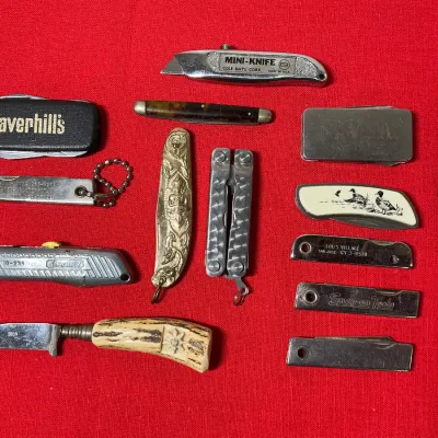 Collection of vintage pocket knives, utility cutters, and advertising novelty blades displayed on a red surface.
