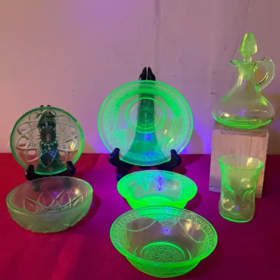 Collection of green uranium glass pieces glowing under blacklight, including a stoppered decanter, small plates, bowls, and a drinking...