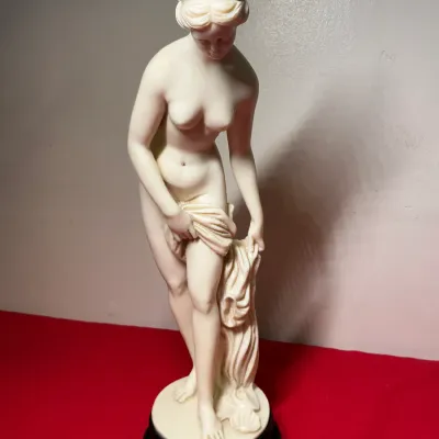 Neoclassical-style ivory resin or composite figurine of a draped female nude, mounted on a dark circular pedestal base.