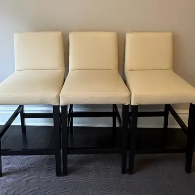 Sale Photo Thumbnail #31: Set of three modern counter stools featuring cream upholstery and dark wood frames with footrests.