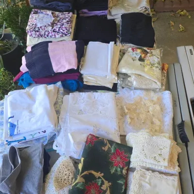 Sale Photo Thumbnail #138: Large assortment of folded clothing, linens, and crochet lace doilies arranged on an outdoor table.