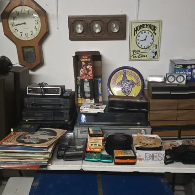 Sale Photo Thumbnail #64: Folding table displaying vintage electronics and media, including a turntable, VCRs, cassette tapes, vinyl records, wall clocks, and a boombox.