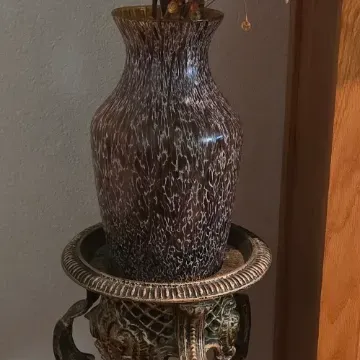 Sale Photo Thumbnail #86: Ornate metal plant stand featuring an intricate floral motif, topped with a dark patterned art glass vase filled with decorative faux botanical sprigs.