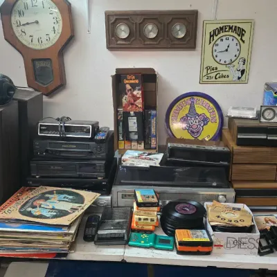 Sale Photo Thumbnail #17: Collection of vintage home audio equipment, media, and decor displayed on a table, including a wall regulator clock, stereo receiver, VHS tapes, vinyl records, and cassette storage drawers.