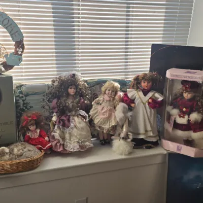 Sale Photo Thumbnail #110: Collection of vintage porcelain and fabric dolls, including one in a display box and a decorative welcome wreath, arranged on a windowsill.