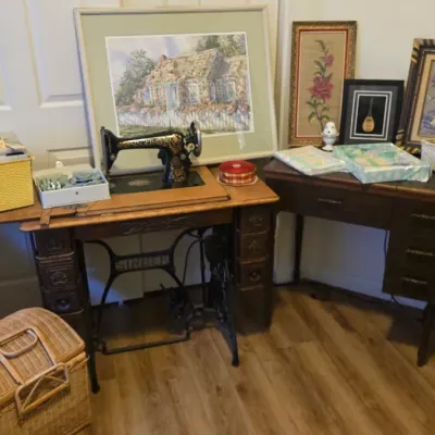 Sale Photo Thumbnail #76: Vintage Singer treadle sewing machine in a wooden cabinet, accompanied by framed artwork, sewing supplies, and a small writing desk.