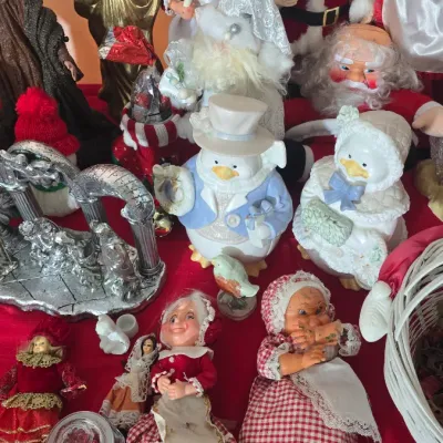 Sale Photo Thumbnail #31: Collection of vintage Christmas figurines and dolls, including Mrs. Claus dolls, Santa figures, dressed duck porcelain statues, and a metallic nativity arch, displayed on a red cloth.