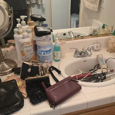 Sale Photo Thumbnail #56: Bathroom counter vanity assortment featuring a metallic makeup mirror, vintage perfumes, evening bags, hair styling tools, and toiletries.