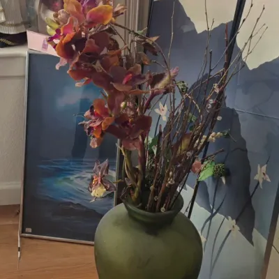 Sale Photo Thumbnail #109: Large olive green floor vase filled with artificial floral stems, set near framed artwork and a boxed porcelain collector doll.