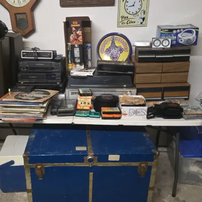 Sale Photo Thumbnail #63: Folding table displaying vintage electronics, vinyl records, VHS tapes, and 8-track cartridges above a blue brass-trimmed steamer trunk.