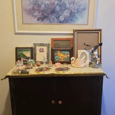 Sale Photo Thumbnail #70: Dark wood two-door cabinet filled with an assortment of collectibles, framed art, and ceramic figurines, with a large framed floral print mounted on the wall above.