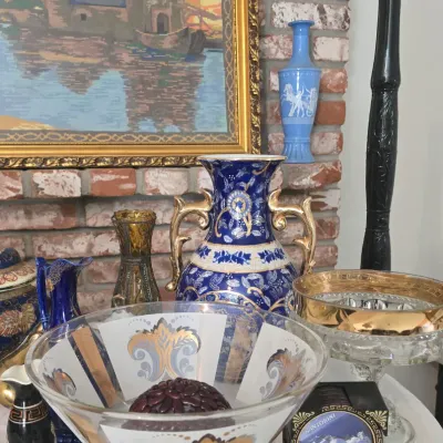 Sale Photo Thumbnail #11: Collection of decorative glassware and ceramics, including a large cobalt blue and gold porcelain vase, a gilt-trimmed glass bowl, and framed artwork displayed on a table against a brick wall.