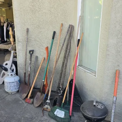 Sale Photo Thumbnail #137: Collection of outdoor garden and yard tools, including various shovels, rakes, an axe, loppers, and a small portable charcoal grill, lined up against an exterior stucco wall.