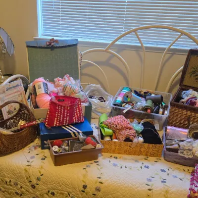 Sale Photo Thumbnail #77: Collection of crafting and sewing supplies, including yarn, ribbon, and embroidery kits displayed on a bed alongside vintage-style baskets and boxes.