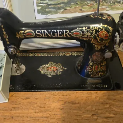 Sale Photo Thumbnail #75: Vintage Singer sewing machine featuring ornate gold decals and a hand wheel, set in a wooden cabinet alongside a box of attachments.