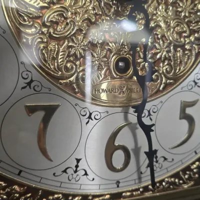 Sale Photo Thumbnail #114: Close-up of a Howard Miller clock face featuring ornate brass filigree detailing, raised Arabic numerals, and decorative black hands.