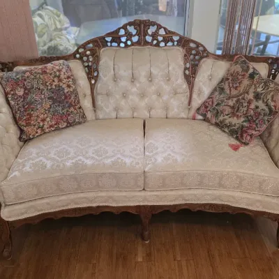 Vintage Victorian-style carved wooden loveseat with cream damask upholstery, tufted backrest, and two floral tapestry throw pillows.
