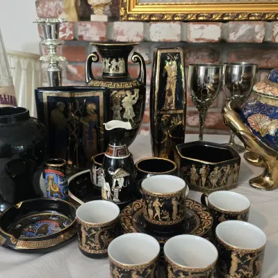Sale Photo Thumbnail #12: Collection of black and gold Greek revival ceramic decor, including a demitasse coffee set, vases, and an ashtray, displayed alongside silver-toned goblets and classical statues.