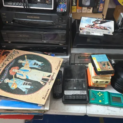 Sale Photo Thumbnail #16: Collection of vintage media and electronics, including vinyl records, cassette tapes, 8-track tapes, a portable cassette player, and stacked VCR and audio components.