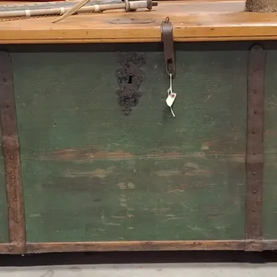 Antique wooden trunk featuring a green-painted body with iron straps and trim, an ornate lock keyhole plate, and a natural wood top.