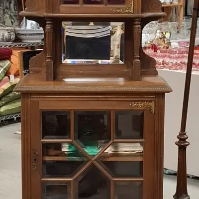 Antique carved wood display cabinet featuring glass doors with geometric mullions, brass corner brackets, a small mirror, and finial...
