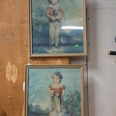 Pair of vintage framed art prints depicting a boy holding a dog and a girl holding a cat, housed in matching thin wooden frames.