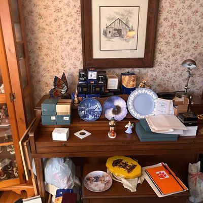 Sale Photo Thumbnail #114: A wooden upright piano displayed with various collectibles, including decorative plates, figurines, boxed items, and framed barn artwork.