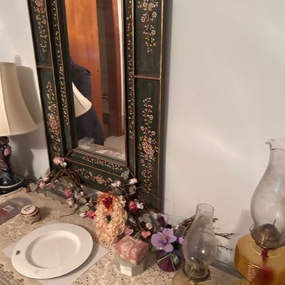Sale Photo Thumbnail #105: Hand-painted floral wood wall mirror framed alongside oil lamps, tabletop decor, and a Limoges book.