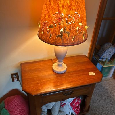 Sale Photo Thumbnail #101: A vintage table lamp with an alabaster base and a pierced floral lampshade, along with a wooden nightstand featuring a single drawer.