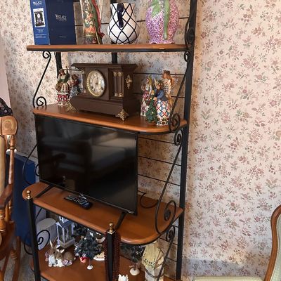 Sale Photo Thumbnail #132: Black wrought iron baker's rack with wooden shelves, holding a small flat-screen television, mantle clock, ceramic pitchers, and decorative figurines.