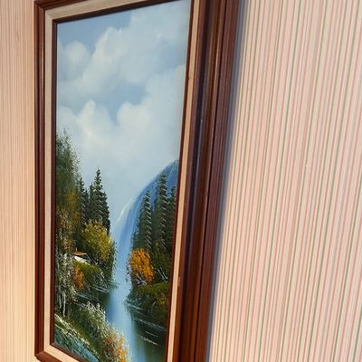Sale Photo Thumbnail #27: Framed vertical oil painting depicting a mountain landscape with a waterfall, pine trees, and a river, housed in a wood frame with cream liner.