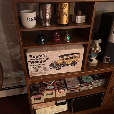 Sale Photo Thumbnail #41: A vintage wooden shelving unit containing drinkware, glass bells, CDs, cassette tapes, and a boxed Jim Beam 1929 Model A Ford "Woodie" decanter.