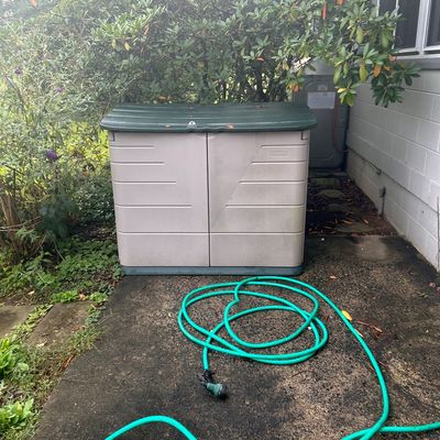 Sale Photo Thumbnail #11: Rubbermaid resin outdoor storage shed with a hinged dark green lid and double front doors, accompanied by a green garden hose.
