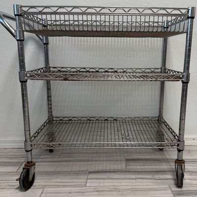 Sale Photo Thumbnail #425: This is a three-tier chrome wire utility rolling cart with a push handle and caster wheels. There is age related wear, including some surface rust. Measurement: 34”L x 18”W x 34”H (approximate). [GG]