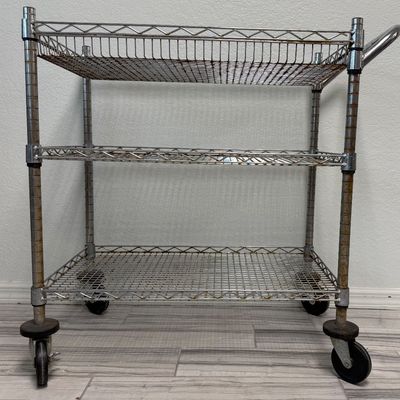 Sale Photo Thumbnail #422: This is a three-tier chrome wire utility rolling cart with a push handle and caster wheels. There is age related wear, including some surface rust. Measurement: 34”L x 18”W x 34”H (approximate). [GG]