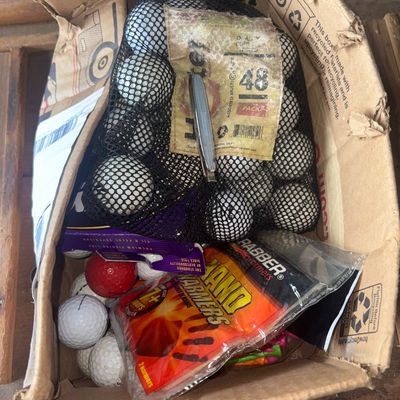 Sale Photo Thumbnail #68: A cardboard box filled with golf supplies and outdoor accessories, including a 48-pack mesh bag of recycled golf balls, loose white and red golf balls, Grabber Hand Warmers packets, fishing line packaging, and a small silver-tone pocket knife. Items show 