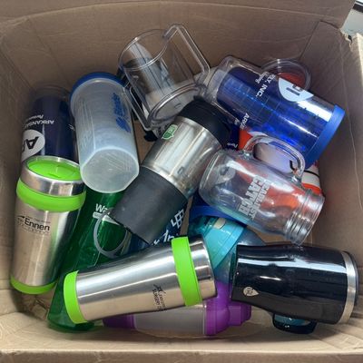 Sale Photo Thumbnail #72: A cardboard box filled with various plastic and stainless steel drinkware. Includes stainless steel travel mugs with green accents, Blender Bottles, clear plastic mugs, insulated tumblers with promotional logos, and water bottles. Items appear to be in us