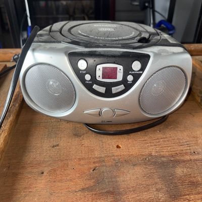 Sale Photo Thumbnail #69: Silver and black Sylvania portable CD player and radio boombox. Features top-loading CD deck, stereo speakers, and front digital display/controls. Includes power cord. Shows surface dust, scuffs, and signs of use.