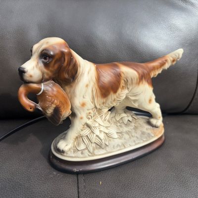 Sale Photo Thumbnail #55: Ceramic or porcelain hunting dog (spaniel or setter type) holding a duck in its mouth, set on a painted base. Detailed hand-painted design in brown, white, and tan tones. Overall good vintage condition with no visible chips or cracks noticed.