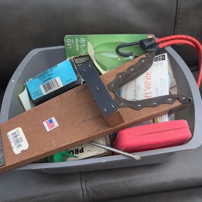 Sale Photo Thumbnail #49: A plastic bin filled with various household items including a wooden shooting or gun rest fixture (Made in USA sticker), a red bungee cord/strap, 40-watt Soft White halogen bulbs in packaging, a small red plastic case, hardware fasteners, and various smal