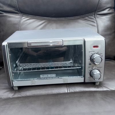 Sale Photo Thumbnail #54: Black+Decker countertop toaster oven with air fry capabilities. Features temperature and timer/toast dial controls, silver-tone finish, glass door, inner wire rack, and mesh air fry basket tray. Appears lightly used with clean interior.
