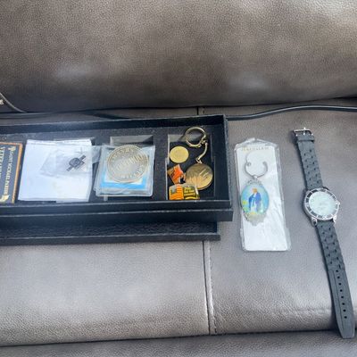 Sale Photo Thumbnail #48: An eclectic collection of small items including a black wristwatch with rubber/silicone strap, a boxed display tray containing a "Thanks A Million" commemorative coin/token, Saint Michael Parish Veterans Network badge, flag pins, keychains including a Jer