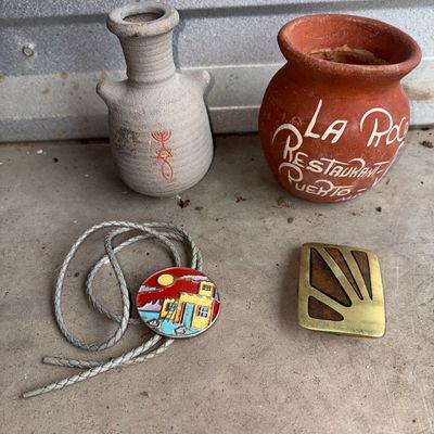 Sale Photo Thumbnail #83: A 4-piece collection of decorative objects including a small grey textured ceramic vase with symbol, a red clay souvenir jar marked 'La Roca Restaurant Puerto...', a Southwestern enamel bolo tie featuring a desert scene with a braided cord, and a brass/go