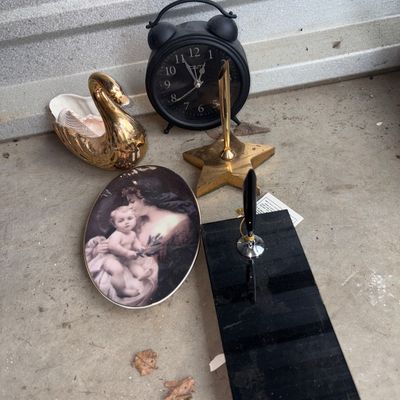Sale Photo Thumbnail #84: An eclectic group lot of decorative items featuring a black matte twin-bell style alarm clock, a gold-tone swan trinket dish holding a shell, a star-shaped brass-tone pen holder, a black stone or acrylic desk pen stand, and an oval portrait print plaque d