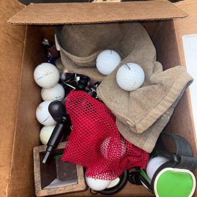 Sale Photo Thumbnail #76: A cardboard box filled with various golf equipment and accessories. Includes multiple golf balls (brands include Bridgestone, Top-Flite, and others), a red mesh ball bag, golf towels, a mini golf bag novelty accessory, and other small items. Items show si