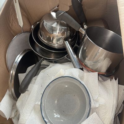 Sale Photo Thumbnail #70: Box lot containing various stainless steel and non-stick pots, saucepans, a fine mesh strainer, metal lids, and ceramic bowl/dishware. Items show normal signs of previous use and handling.