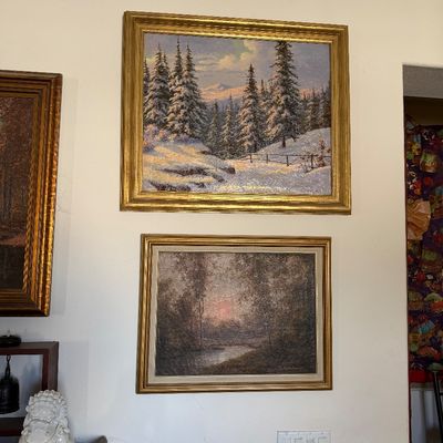Sale Photo Thumbnail #12: Two framed landscape oil paintings, including a snowy mountain forest scene above a moody sunset woodland river scene, both housed in ornate gold frames.