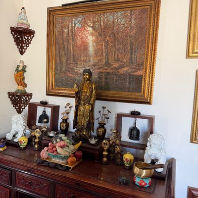 Sale Photo Thumbnail #9: Asian alter display featuring a standing gilded Buddha statue, a carved jade fruit bowl, white foo dogs, hanging bells, and a large framed autumn forest painting.
